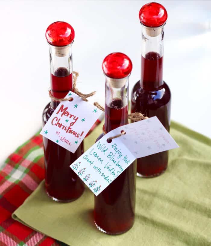 three tall glass bottles with red tops filled with blueberry infused vodka with tags tied on them that say "Merry Christmas"