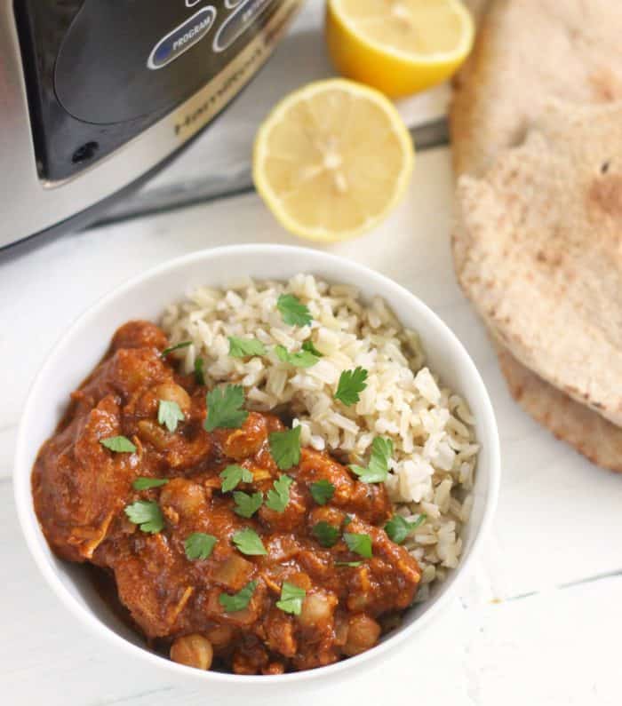 white bowl with chicken tikka masala and brown rice topped with cilantro, naan, lemon