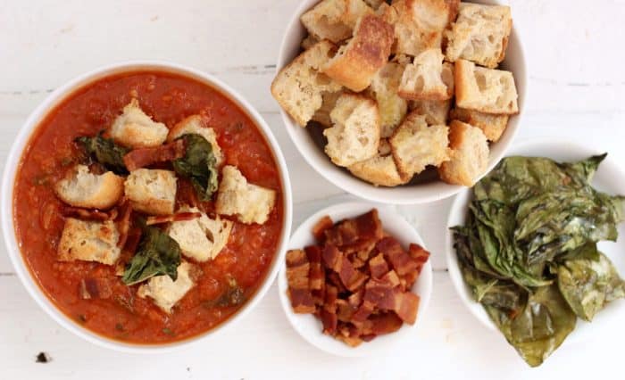 white bowls with homemade croutons, crispy basil, cooked bacon, and tomato soup covered in toppings
