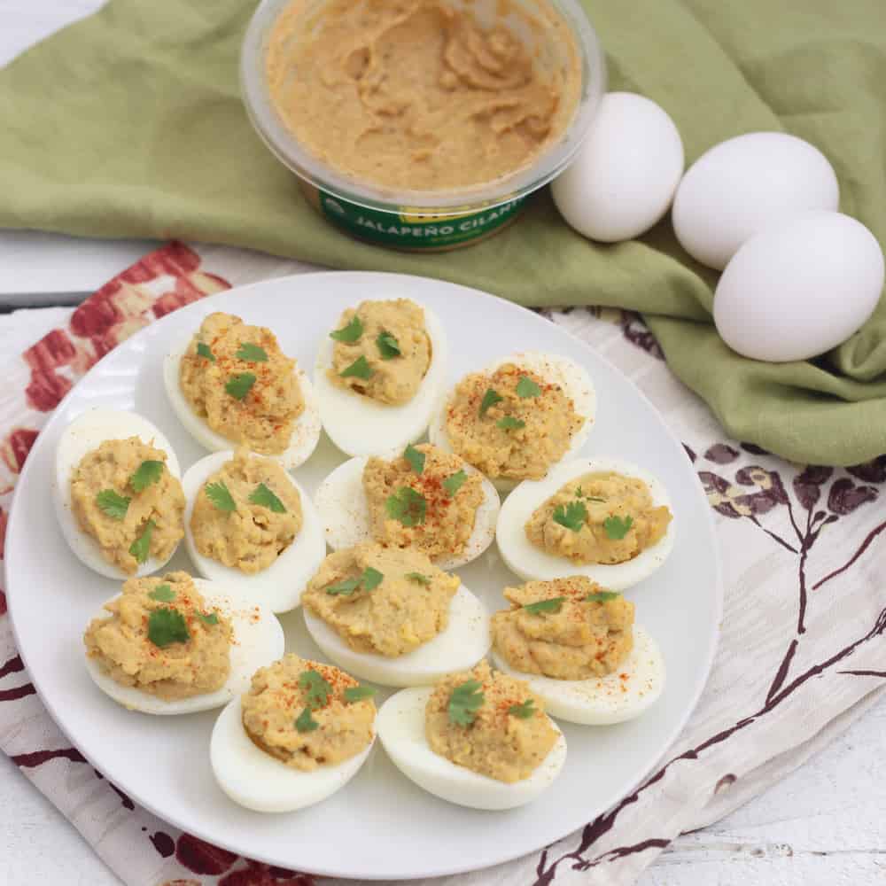 white plate on floral napkin with deviled eggs, green napkin topped with hard boiled eggs and container of hummus