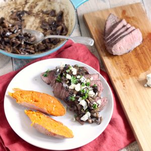 red napkin topped with white plate that has open sweet potato, steak covered in mushroom blue cheese sauce and parsley, cutting board with steak and skillet with sauce