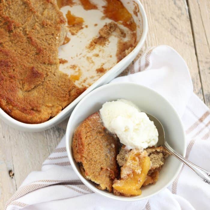 white bowl of peach cobbler with ice cream and spoon on towel with baking dish of cobbler