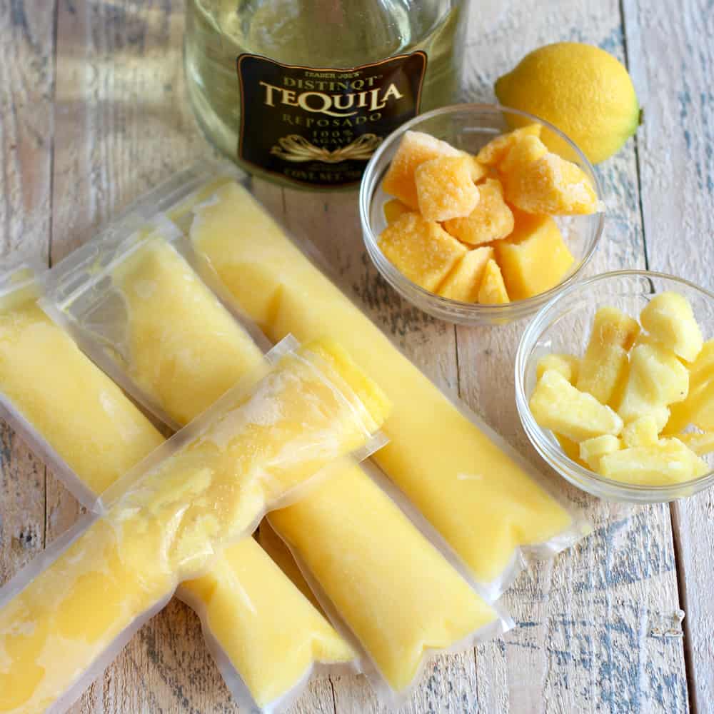 homemade Mango Pineapple Popsicles, bottle of tequila, lemon, and clear bowls with frozen mango and pineapple on a wooden table
