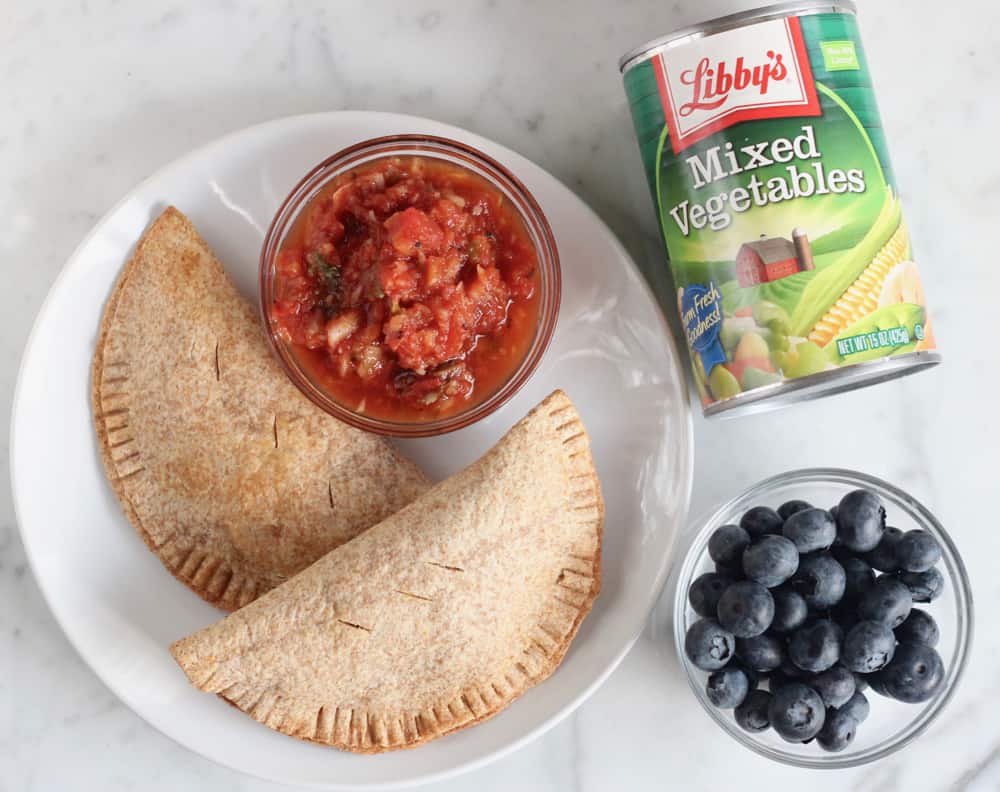 a white plate with a bowl of salsa and two Turkey and Vegetable Empanadas next to a can of mixed vegetables and a bowl of blueberries.