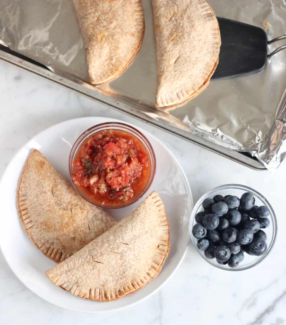 two turkey and vegetable empanadas on a plate with a bowl of salsa next to a bowl of blueberries and a baking sheet with extra empandas.