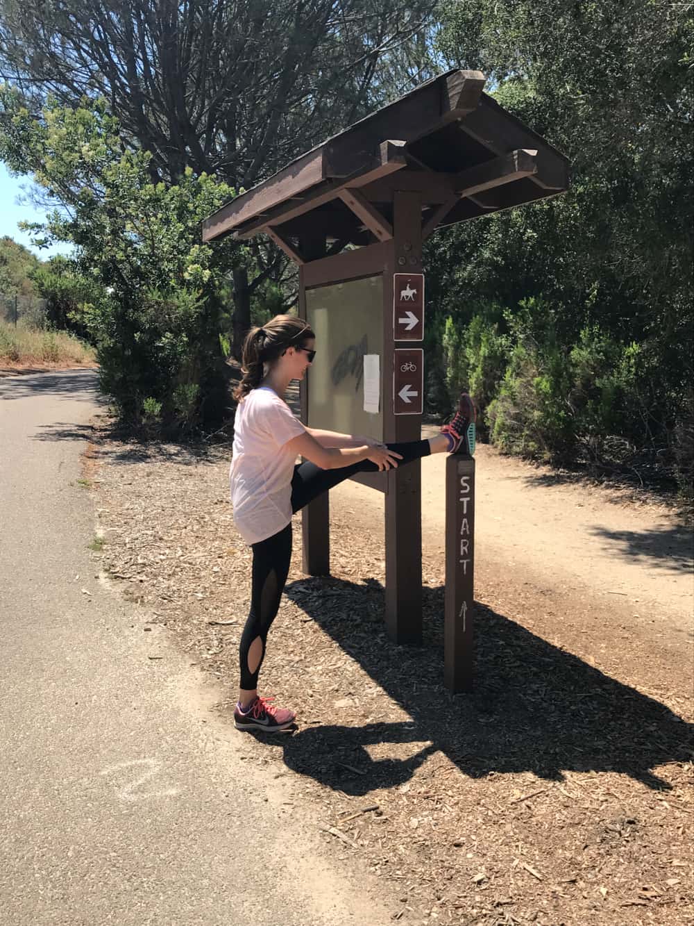 brunette female stretching leg on a park sign near a hiking trail map