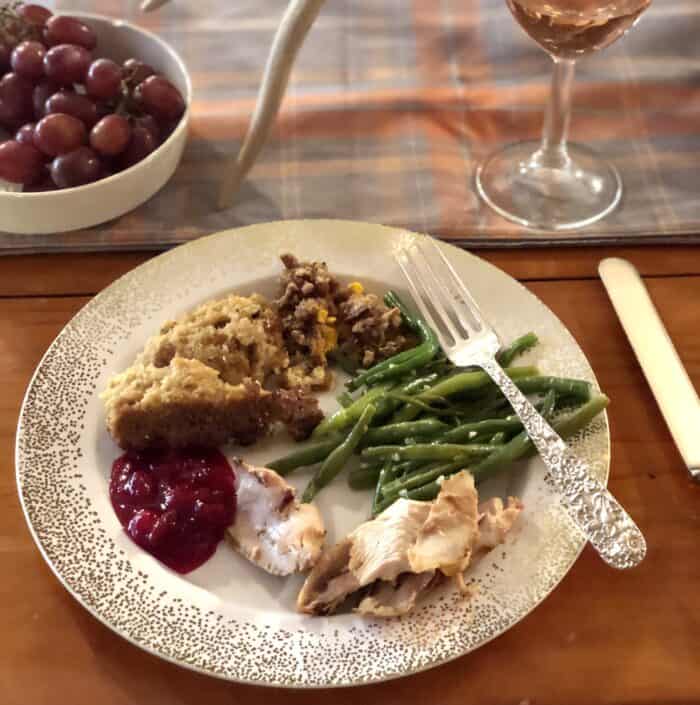 white plate covered in gold flecks with turkey, cranberry sauce, dressing, sweet potato casserole, green beans and silver fork with wine and grapes on table
