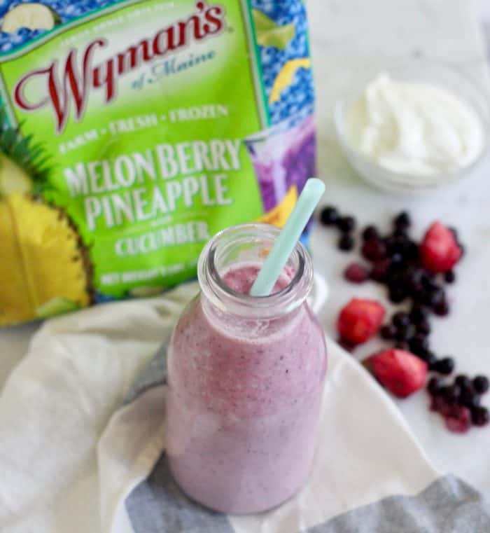 light purple smoothie in clear glass with a blue straw, and yogurt, berries, and frozen bag of melon berry pineapple