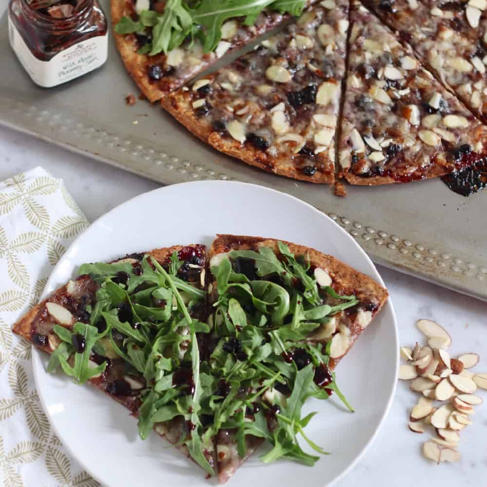 white plate with two slices of arugula topped blueberry gruyere pizza next to sliced almonds and a baking sheet with extra pizza and blueberry jam.