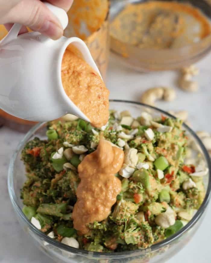 pouring vegan cashew red bell pepper cheese sauce over vegan broccoli salad