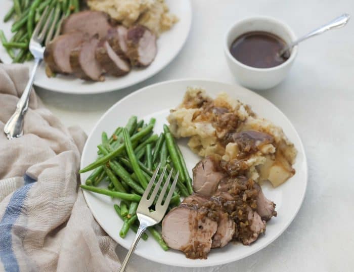 white plates with pork tenderloin, green beans, and mashed potatoes with fork, gravy, tan and blue napkin