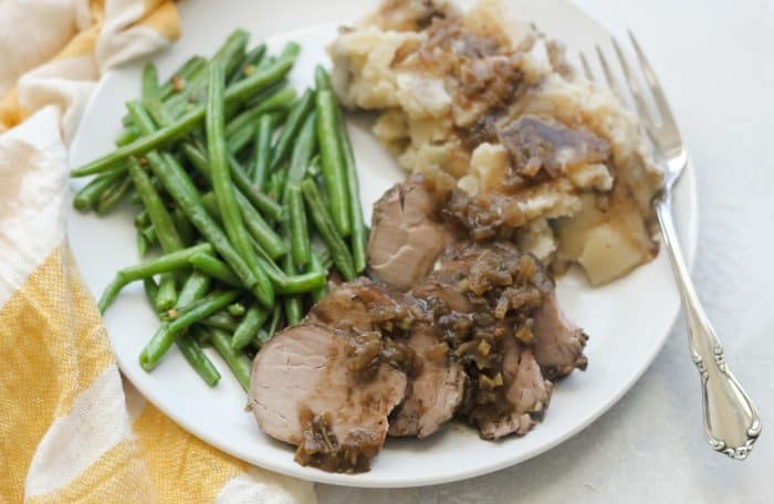 pork tenderloin with gravy, mashed potatoes, and green beans on white plate with yellow and white striped napkin