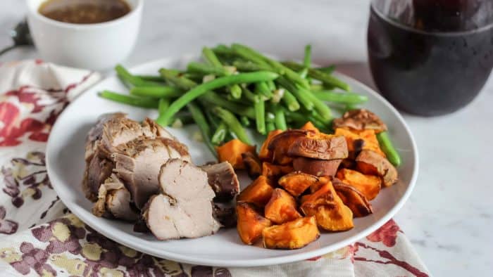 glass of wine, bowl of gravy, white plate on a flower napkin with pork tenderloin, roasted sweet potatoes, and green beans