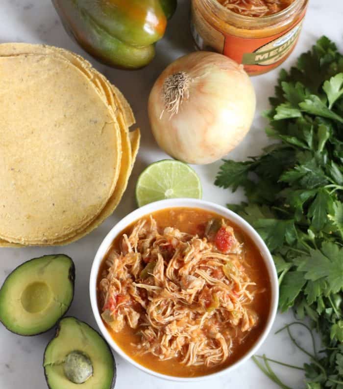 bowl of salsa chicken with tortillas, avocados, lime, onion, green bell pepper, jar of salsa, and parsley