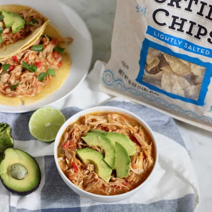 bowl of salsa chicken with sliced avocados, bag of tortilla chips, tacos on the side