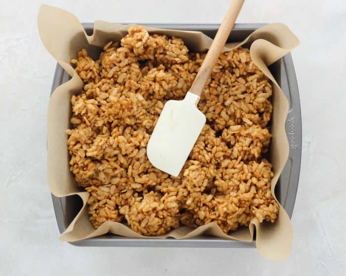 rice crispy treats being pressed into baking dish with parchment paper by a spatula