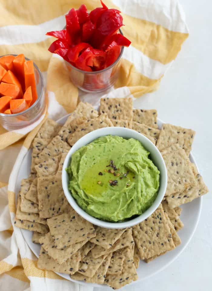 yellow and white striped towel with sliced carrots and bell peppers and a plate of crackers with bowl of spinach avocado hummus