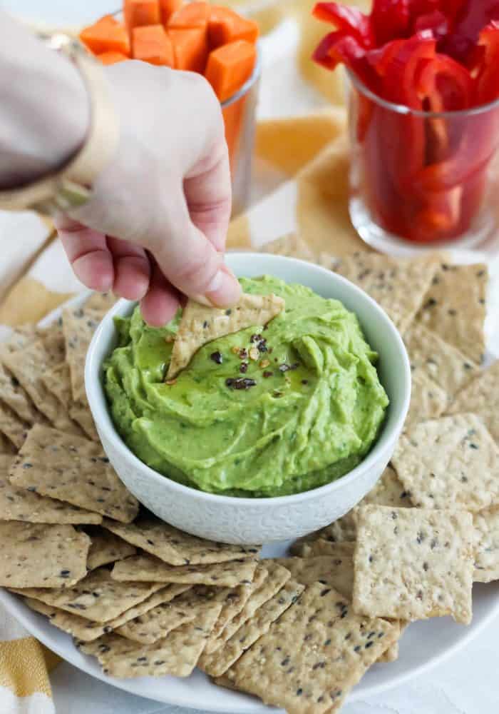 hand dipping a cracker into bowl of avocado hummus on plate with crackers, sliced bell peppers and carrots