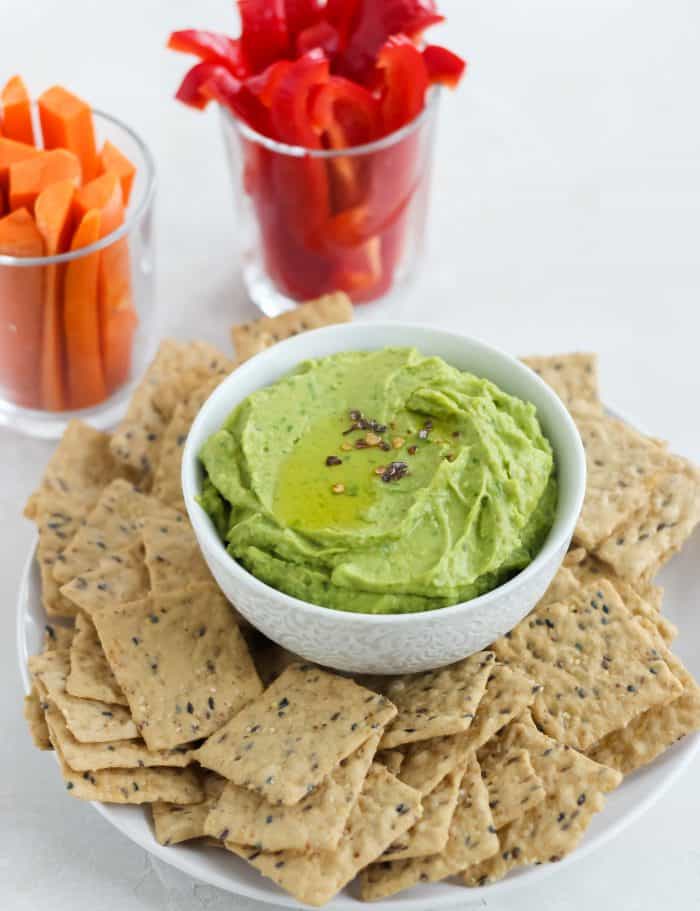 plate of crackers with white bowl of avocado spinach hummus, glasses full of sliced carrots and bell peppers