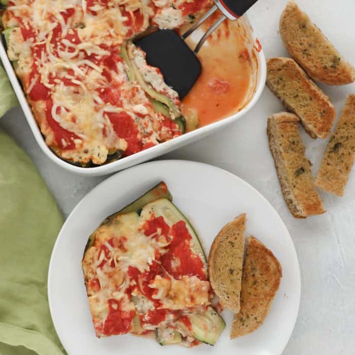 zucchini lasagna on white plate with garlic bread and casserole dish with spatula