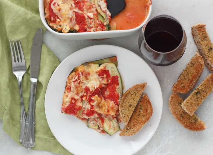 zucchini lasagna on white plate with garlic bread and casserole dish, red wine