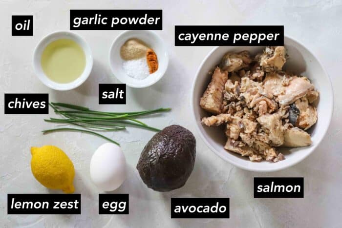 ingredient photo with black text labeling item: oil, garlic powder, cayenne pepper, salt, canned salmon in a white bowl, avocado, egg, lemon, chives
