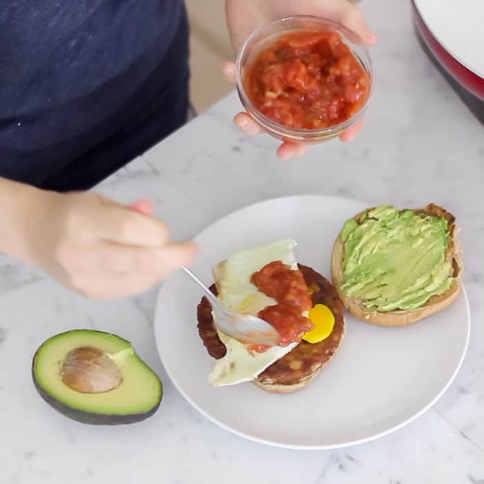 breakfast burger with fried egg and avocado being topped with salsa on white plate