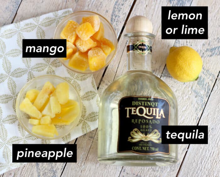 bowls of frozen chopped mango and pineapple with a lemon and a bottle of tequila on a wooden table