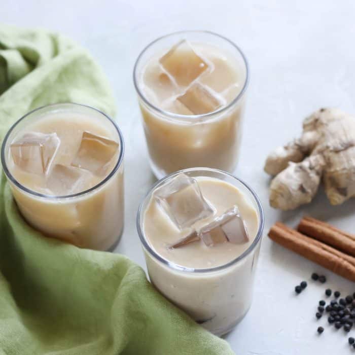 three small glasses of iced chai lattes with fresh ginger, cinnamon sticks, black peppercorns