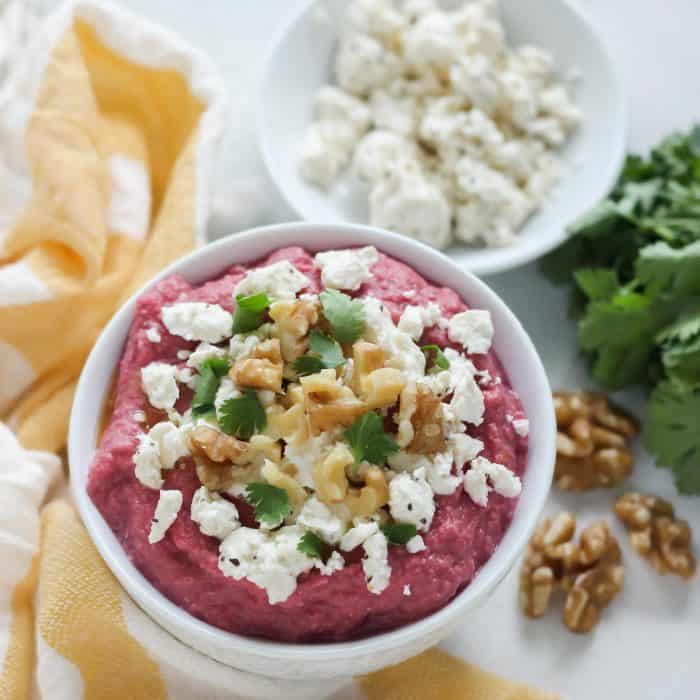 bowl of beet hummus with toppings and bowl of feta, walnuts, cilantro, yellow and white towel