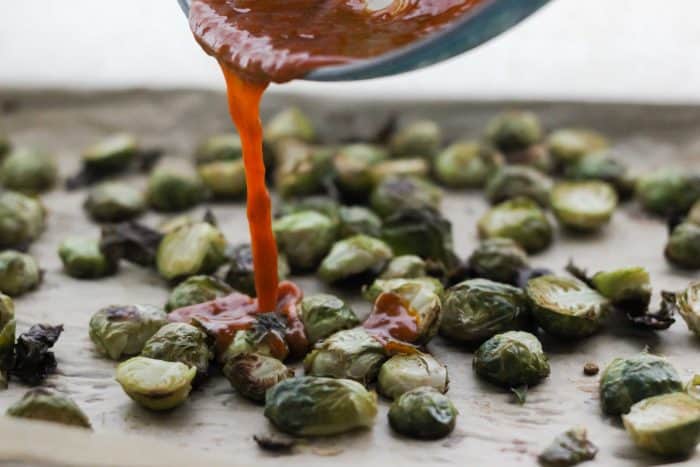 butter-buffalo sauce pouring onto roasted Brussels sprouts on parchment lined baking sheet