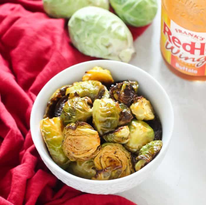 red towel with white bowl of roasted Brussels sprouts with buffalo sauce and fresh Brussels sprouts