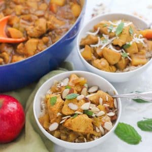 two white bowls of chicken apple curry topped with sliced almonds and fresh mint, apple, and blue pot with more chicken curry sauce