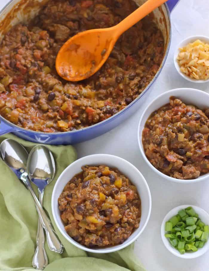 two white bowls of pumpkin chili with a blue pot of pumpkin chili, green onions, cheddar cheese, spoons, green napkin