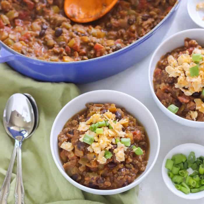 green napkin with spoons, two white bowls with pumpkin chili topped with cheese and green onions, blue pot of chili with orange spoon