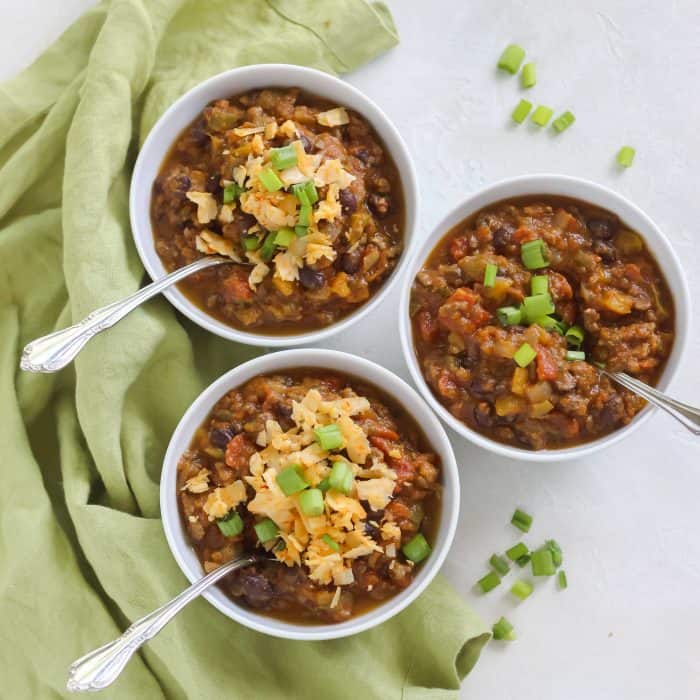 three white bowls of pumpkin chili on a green towel with green onions and cheese