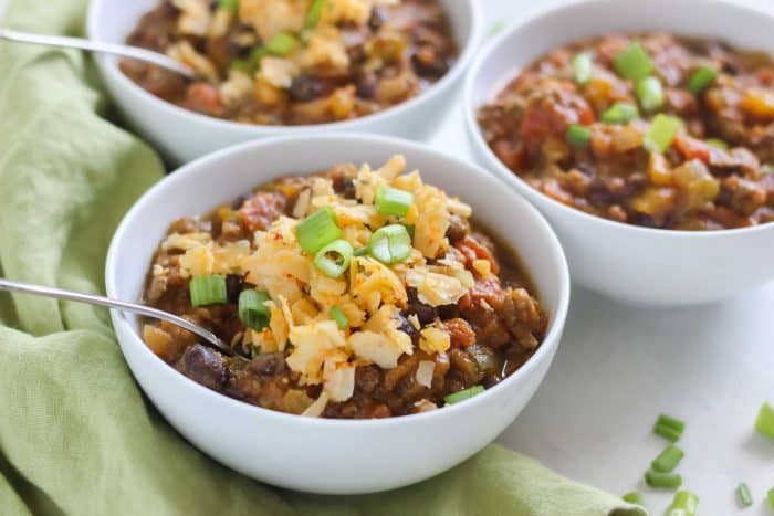 three white bowls of pumpkin chili topped with cheese and green onions with spoons on green napkin 