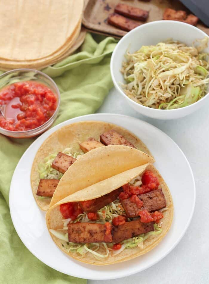 tofu tacos on a white plate with green napkin and bowl of salsa, tortillas, slaw, and tofu