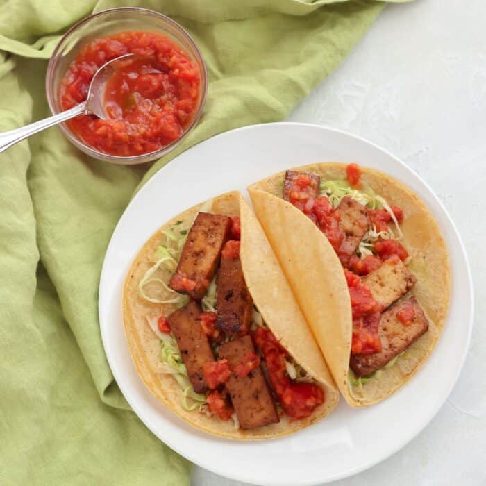 small bowl of salsa on green napkin with plate of two tacos with tofu and slaw