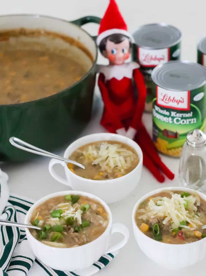 three white cups of cheeseburger chowder with spoons, canned veggies, elf on the shelf, pot of soup