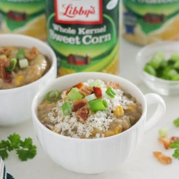 white cup of cheeseburger chowder topped with cheese, bacon and green onions with another cup of soup and canned veggies