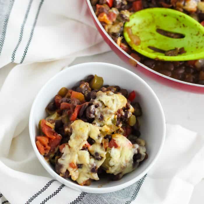 bean casserole in a white bowl with the casserole in a skillet on the side and a green slotted spoon