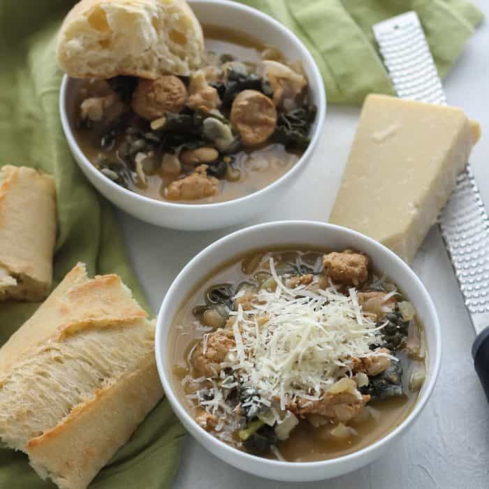 two bowls of sausage kale white bean soup topped with parmesan cheese, with french bread and fresh kale