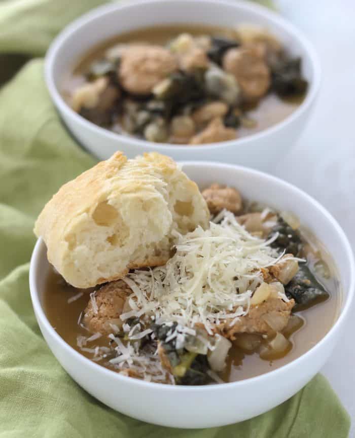two white bowls of white bean sausage kale soup topped with parmesan cheese and slice of bread