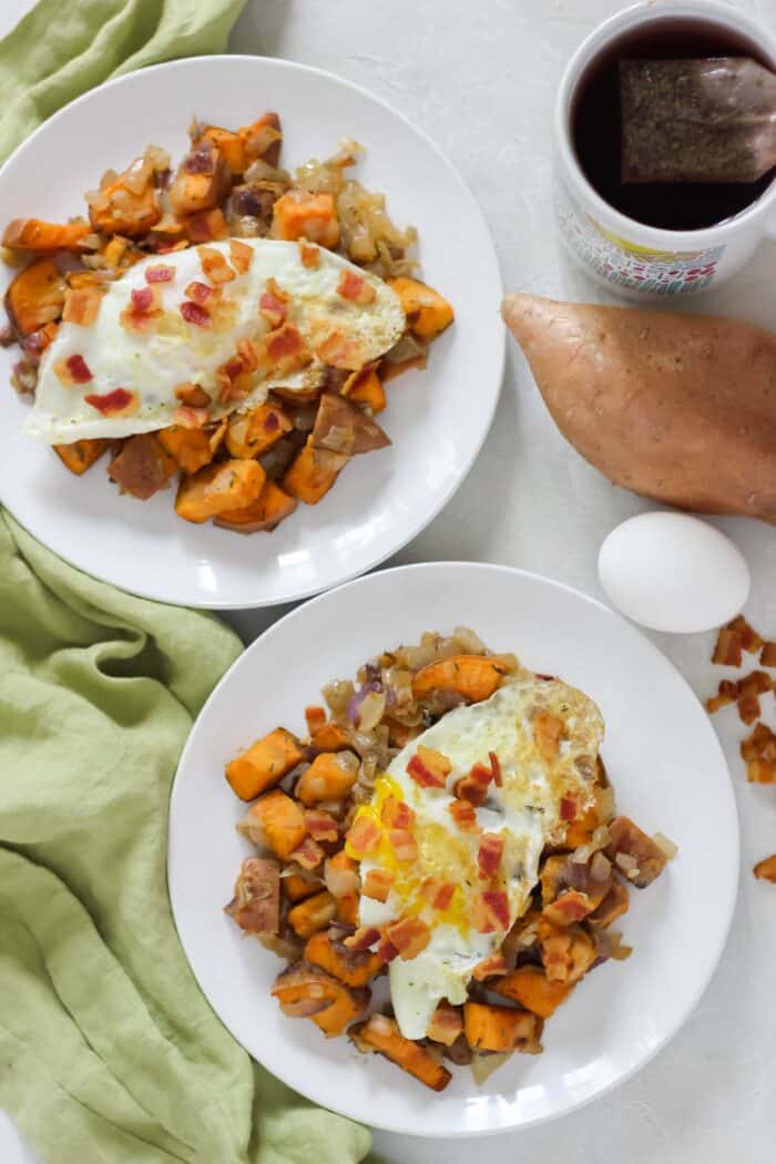 white plate on white countertop with sweet potato and bacon hash topped with fried eggs and bacon crumbles with sweet potato, egg, cup of tea, and bacon