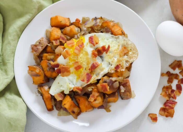 white plate of sweet potato hash with fried egg and bacon on top sitting on a green napkin on white counter with one fresh egg and bacon crumbles