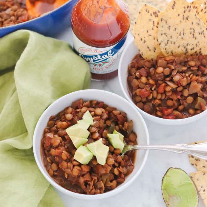 two white bowls of chili, one with avocado and a spoon, the other with tortilla chips popping out. sitting on a green napkin with hot sauce and a pot of chili