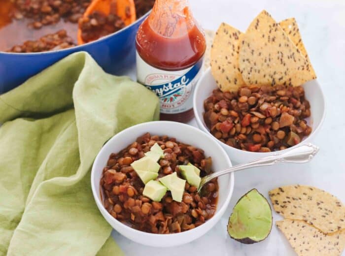 two white bowls of chili one with avocado, the other with chips. a green napkin and a blue pot of lentils with avocado and chips and hot sauce