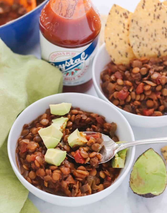 spoon in a white bowl of chili with avocados, green napkin on white counter with avocado peel, another cup of chili with chips, and hot sauce