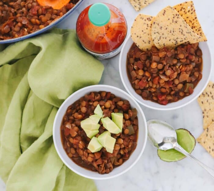 overhead look at two bowls of chili with avocado and chips on a green napkin with a pot of chili and hot sauce